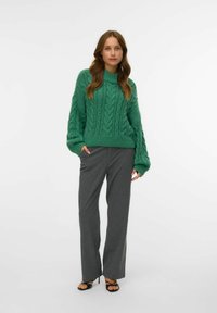 Frau trägt einen grünen Kabelstrickpullover, graue Nadelstreifenhose und schwarze Sandalen mit offenen Zehen, steht vor einem schlichten weißen Hintergrund.