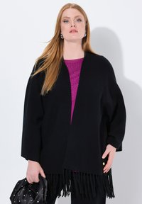 Schwarzer, gestrickter Wickel mit fransigem Saum, der eine offene Vorderseite und strukturierte Ärmel hat. Darunter ein lila Top, das mit Glitzer verziert ist.