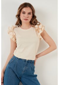 Beige top s krátkým rukávem a volány na ramenech, spárovaný s vysokými modrými džíny. Hladká textura a uvolněný střih.