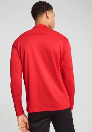 Giovane uomo di spalle, indossa una maglia sportiva rossa a maniche lunghe e pantaloni neri su uno sfondo chiaro e uniforme.