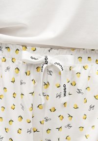 Κοντινή λήψη λευκού υφασμάτινου παντελονιού με κίτρινα σχέδια λεμονιών και μαύρο κείμενο "Hugo", με λευκό κορδόνι δεμένο σε φιόγκο με λογότυπο "HUGO".