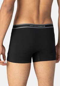 Homme portant un boxer noir avec une ceinture grise portant le texte "REGARDE TOUJOURS DU BON CÔTÉ", vu de derrière sur un fond blanc uni.
