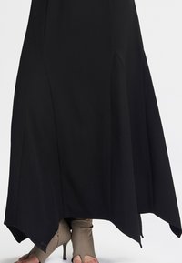 Maxi abito nero con una silhouette fluida, caratterizzato da un orlo inclinato e cuciture laterali. Stivali con tacco aperti beige visibili sotto l'abito.