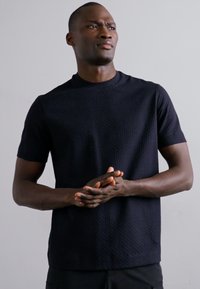 Μαύρη κοντομάνικη μπλούζα με υφή, στρογγυλή λαιμόκοψη και διακριτό γεωμετρικό μοτίβο, κατασκευασμένη από ελαφρύ ύφασμα.