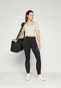 Noor naine mustades retuusides, beežis lühikeses topis ja valgetes tossudes, hoiab musta spordikotti õlal, seistes ühetaolise taustaga.