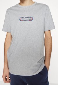 Camiseta de algodón gris con cuello redondo, que presenta un logo azul, rojo y gris que dice "HILFIGER NEW YORK CITY 1985" en el pecho.