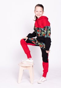 Czerwono-czarny dres z nadrukiem kwiatowym z zielonymi liśćmi i papugami. Bluza z kapturem kontrastuje z jednokolorowymi spodniami.