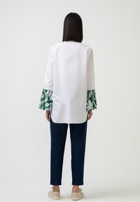 Blusa bianca con polsini a motivo floreale verde, maniche lunghe e taglio rilassato. Abbinata a pantaloni in denim blu scuro e scarpe beige.