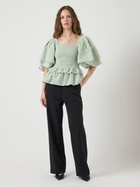 Donna che indossa una blusa verde a quadri con maniche a sbuffo, corpetto arricciato e orlo a balze, abbinata a pantaloni neri a gamba larga e scarpe nere.