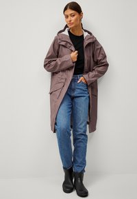Gabardina larga de un suave color malva con capucha, bolsillos con cremallera y una textura lisa, combinada con unos vaqueros oscuros y botines negros.
