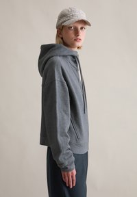 Grijze capuchontrui met een rits aan de voorkant, gestructureerde stof en verstelbare trekkoorden, gecombineerd met een donkere broek en een geblokte pet.