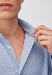 Close-up of a person adjusting the collar of a light blue textured button-up shirt, showing neck and lower face.