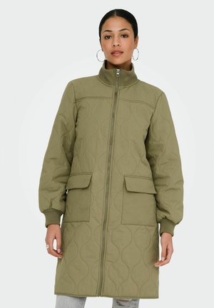 Femme portant un manteau matelassé mi-long vert olive avec une fermeture éclair frontale, des poches à rabat, des poignets côtelés et des boucles d'oreilles en anneau, debout devant un fond blanc.