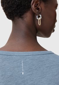 Grå-blå T-shirt med en afslappet halsudskæring, prydet med et logo trykt på ryggen. Guld-tone kædeøreringe med geometriske accenter bæres.