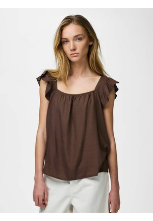 Kobieta z długimi blond włosami, ubrana w brązową, bezrękawnikową, marszczoną bluzkę oraz białe spodnie na jasnym, jednolitym tle.