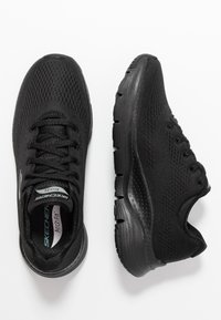 Zapatillas deportivas negras con parte superior de malla transpirable, superficie texturizada y suela acolchada. Las características incluyen cordones y un logo en la lengüeta.