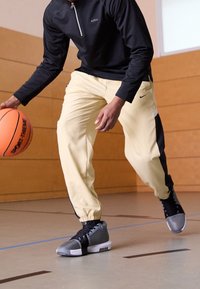 Schwarzes Langarmshirt mit Reißverschluss, gelbe Sporthose mit schwarzen Akzenten und graue Basketballschuhe auf einem Sportboden mit einem Basketball.