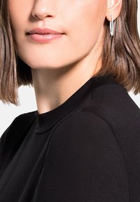 Zwart hoogneck top met een gladde textuur, met een getailleerd ontwerp. Zilveren ringoorbel accentueert de look. Korte, steile haren.