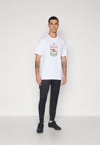 Wit T-shirt met een kleurrijk Arsenal-logo op de voorkant, gecombineerd met zwarte sportbroek met witte zijstrepen en zwarte sportschoenen.