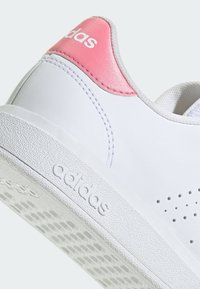 Zapatilla blanca hecha de material sintético con un acento de talón rosa. Suela texturizada con perforaciones. Logo mostrado en el lateral.