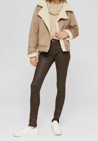 Brun fuskpälsjacka med krämfärgad krage, kombinerad med åtsittande chokladbruna byxor och vita sneakers. Mjuk textur, lager-på-lager-design.