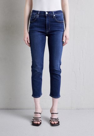 Person wearing cropped dark blue jeans and black strappy sandals, standing against a light gray textured wall.