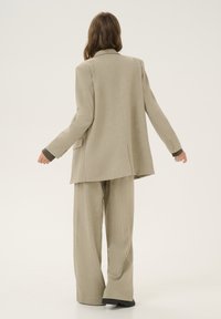 Beige skræddersyet blazer med en enkelt ventil, parret med brede bukser. Struktureret stof, kontrasterende mørke manchetter og knapdetaljer på ærmerne.