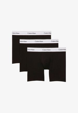 Jonge man met kort donker haar staat blootsvoets tegen een grijze achtergrond, draagt zwarte boxershorts met een witte Calvin Klein tailleband.