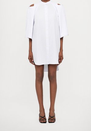 Modella che indossa un vestito bianco con bottoni, spalle scoperte e maniche tre quarti, abbinato a sandali neri con tacco alto e cinturini.