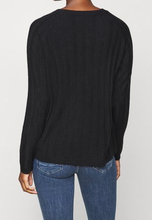 Person iført en sort ribstrikket langærmet sweater og blå denimjeans, set bagfra mod en neutral baggrund.