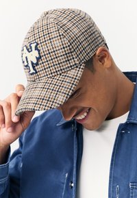 Braun-beige kariertes Käppi mit blauem Sticklogo, gebogener Krempe und strukturiertem Wollmaterial. Leicht nach vorne geneigt getragen.