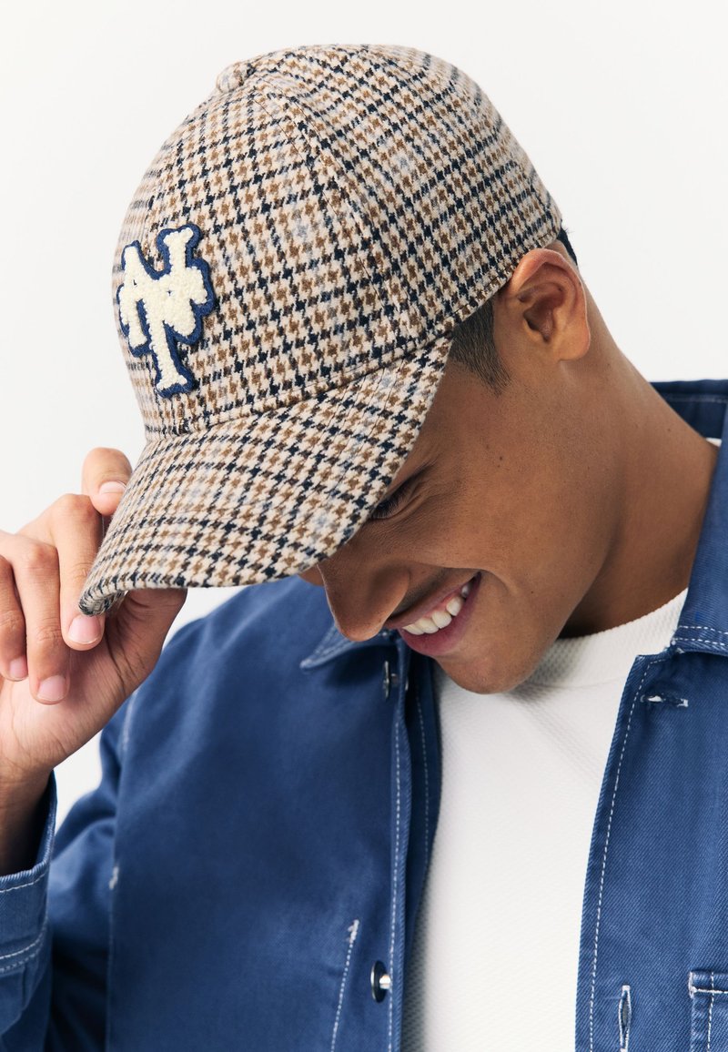 Braun-beige kariertes Käppi mit blauem Sticklogo, gebogener Krempe und strukturiertem Wollmaterial. Leicht nach vorne geneigt getragen.
