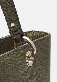Närbild av en texturerad olivgrön handväska rem fäst med en gyllene ring och nitar, med GUESS-märkesgravyr på metallhardwaren.