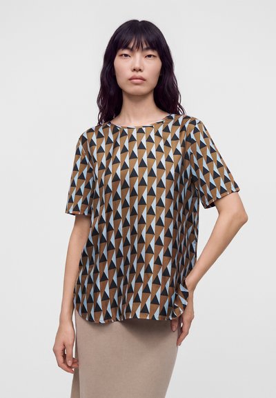 Femme portant une chemise à manches courtes avec des motifs triangulaires géométriques marron, noir et bleu clair et une jupe beige sur un fond uni.