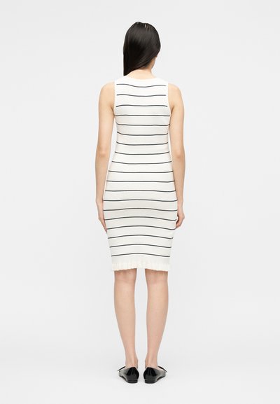 Γυναίκα που στέκεται με την πλάτη, φοράει ένα άσπρο φόρεμα χωρίς μανίκια μέχρι το γόνατο με λεπτές μαύρες οριζόντιες ρίγες και μαύρα επίπεδα παπούτσια.