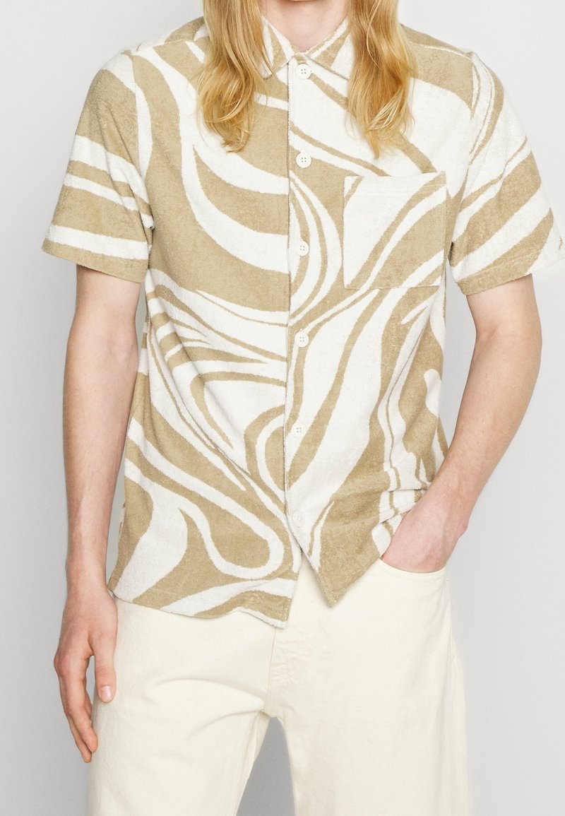 Personne portant une chemise beige et blanche à manches courtes avec des motifs abstraits en spirale et un pantalon couleur crème, une main dans la poche.