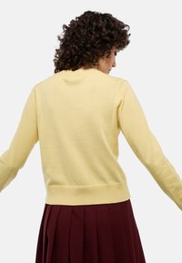 Pull en tricot jaune à manches longues, avec un col et un ourlet côtelés. Texture lisse, ajusté à la taille, montré de dos sur un fond neutre.