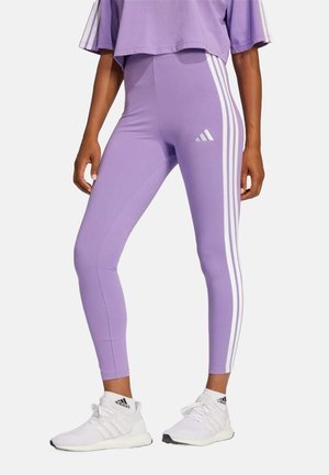Leggings lilla con accenti a tre strisce bianche sui lati e logo Adidas. Il materiale sembra elastico e aderente. Scarpe da ginnastica bianche.