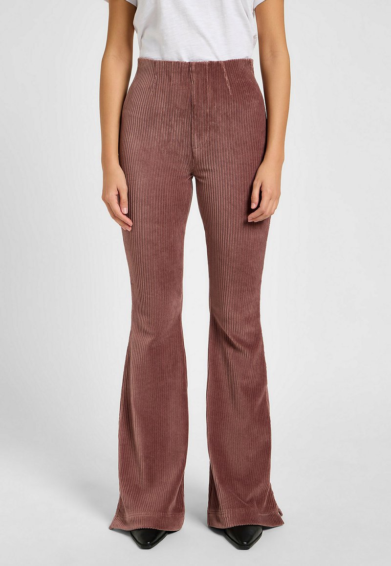 Pantaloni a zampa d'elefante in corduroy marrone con righe verticali, vita alta e orlo svasato, abbinati a una semplice maglietta bianca e scarpe nere.