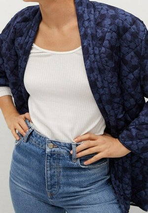 Persoon draagt een wit geribd tanktopje, blauwe jeans met hoge taille en een donkerblauwe gebloemde doorgestikte jas met de handen op de heupen.