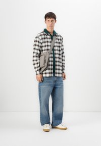 Ung mand iført ternet skjorte, teal polo, vidde jeans, hvide sneakers og grå crossover-taske, der står mod en hvid baggrund.