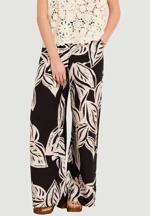 Woman wearing black wide-leg pants with large beige leaf patterns, a beige crochet top, and tan platform sandals.