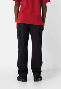 Zwarte cargobroek met een relaxte pasvorm, voorzien van zijzakken en een gladde stoftextuur, gecombineerd met een rood T-shirt met korte mouwen.