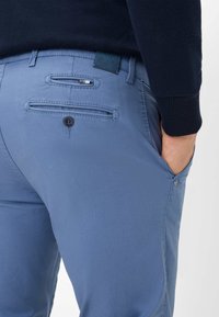 Lichtblauwe broek van een gestructureerde stof, met twee achterzakken met kleppen en knopen, en een klein leren label met merknaam op de tailleband.