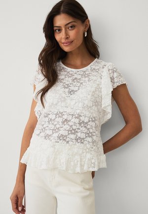 Vrouw met lang bruin haar die een witte, bloemige, kanten top met korte mouwen en crèmekleurige broek draagt, staand tegen een effen lichte muur.