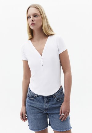 Jeune femme aux longs cheveux blonds portant un haut blanc à manches courtes avec col en V boutonné et un short en jean bleu.