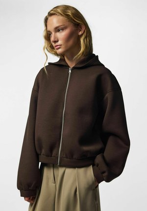 Jeune femme aux cheveux blonds ondulés portant un sweat à capuche marron foncé zippé et un pantalon beige, regardant de côté sur un fond blanc uni.