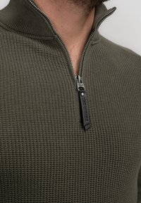 Olivgrüner, strukturiertes Zip-up-Pullover mit hohem Kragen und silbernem Reißverschluss. Das Material wirkt gestrickt und verleiht einen gemütlichen Look.