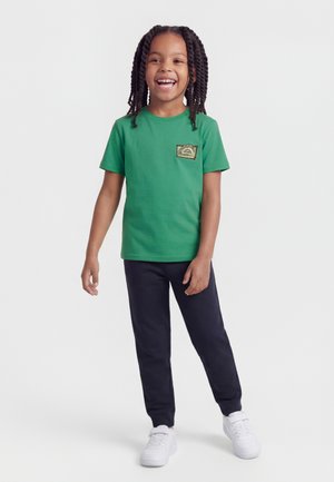 Enfant souriant aux cheveux tressés portant un t-shirt vert avec un écusson, un pantalon noir et des baskets blanches, debout devant un fond uni.