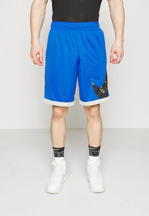 Homme portant un short de sport bleu avec un liseré blanc, des chaussettes noires à motifs et des baskets blanches, debout devant un fond uni.